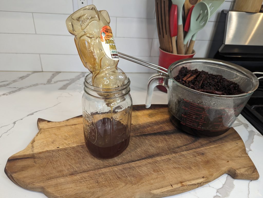 draining honey into mason jar on wood cutting board with elderberry syrup straining into measuring cup