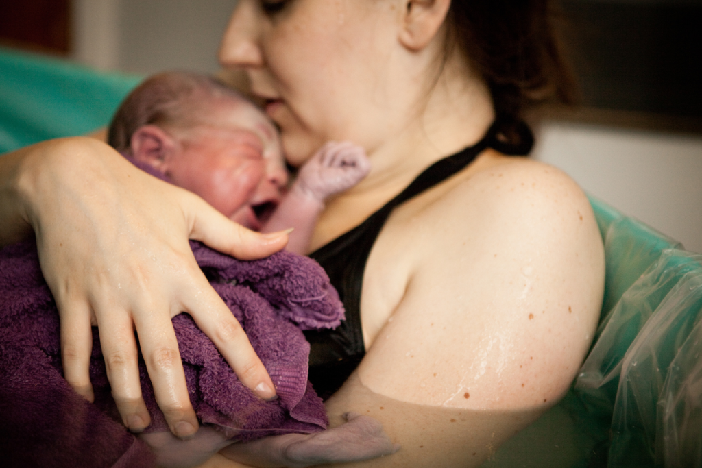 woman in birthing tub with newborn baby in her arms in purple towel