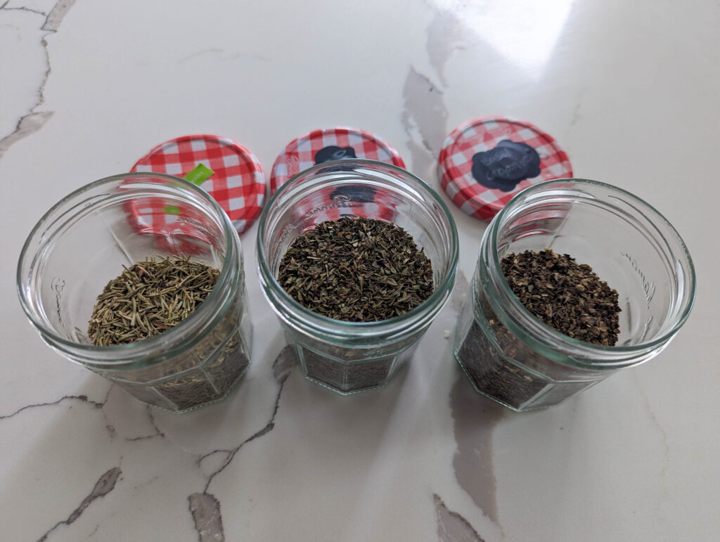 dried herb sof oregano, rosemary and basil in glass jars on white counter