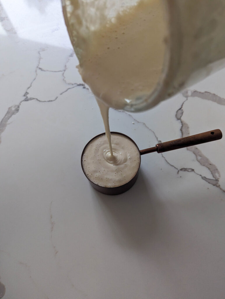 pouring sourdough starter into a copper measuring cup on white counter