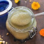 spooning up the calendula & chamomile body scrub with flowers on wooden cutting board